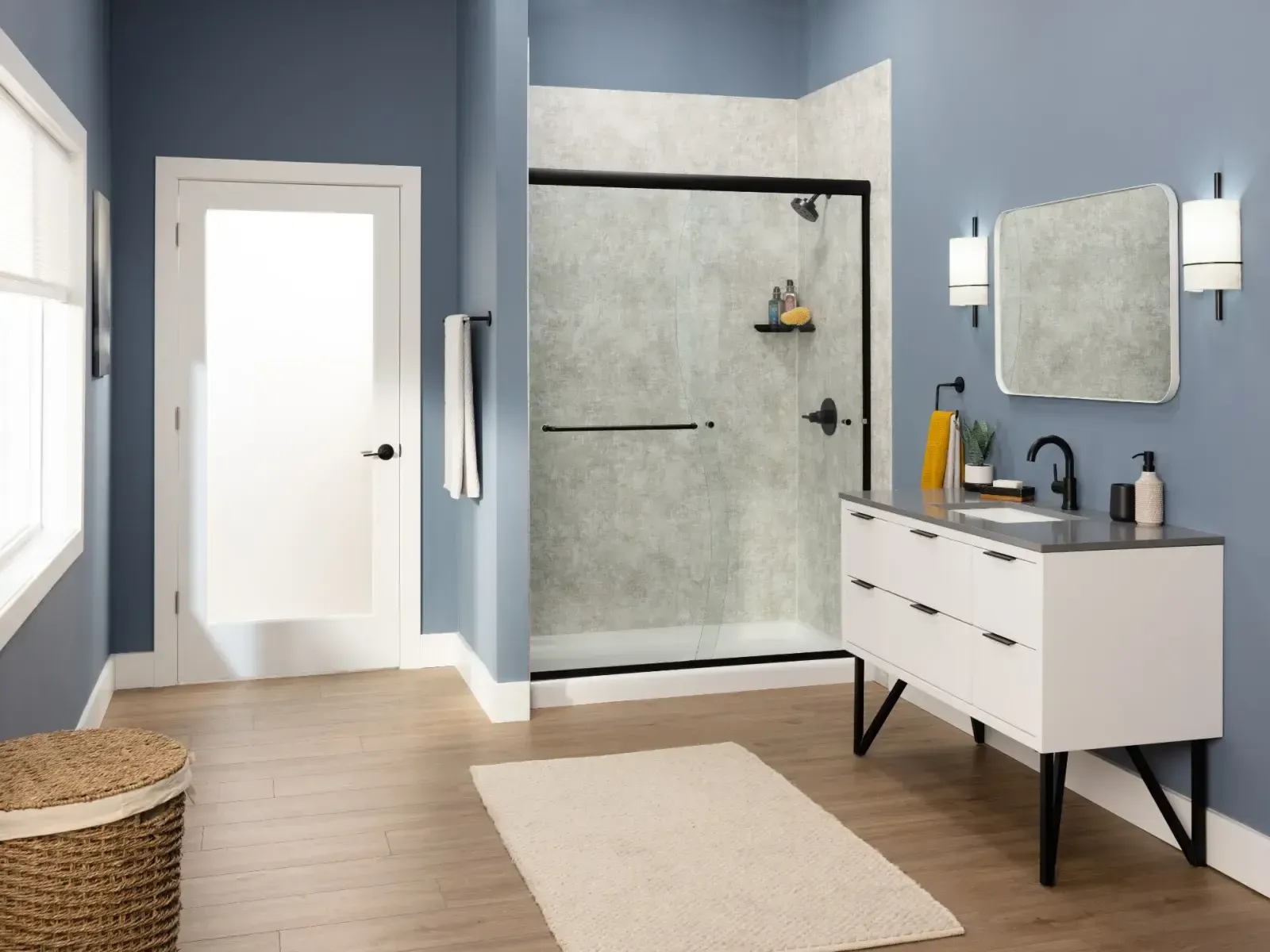 Modern bathroom with blue walls, featuring a glass shower with black trim, white vanity with mirror, wicker basket, and wooden floor.