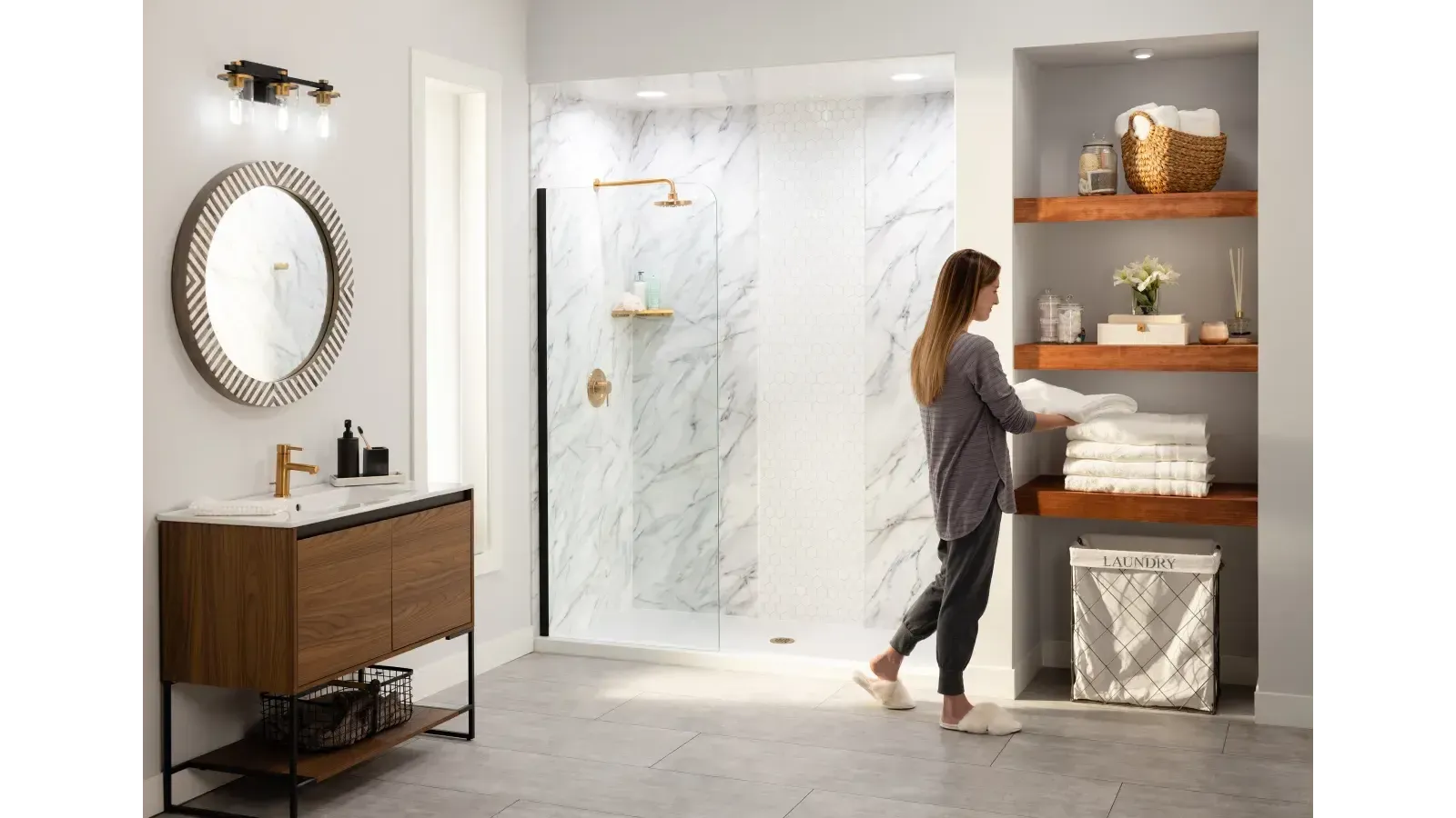 A woman arranges towels on wooden shelves in a modern bathroom with a marble shower, a circular mirror, and a wooden vanity, creating a serene atmosphere.