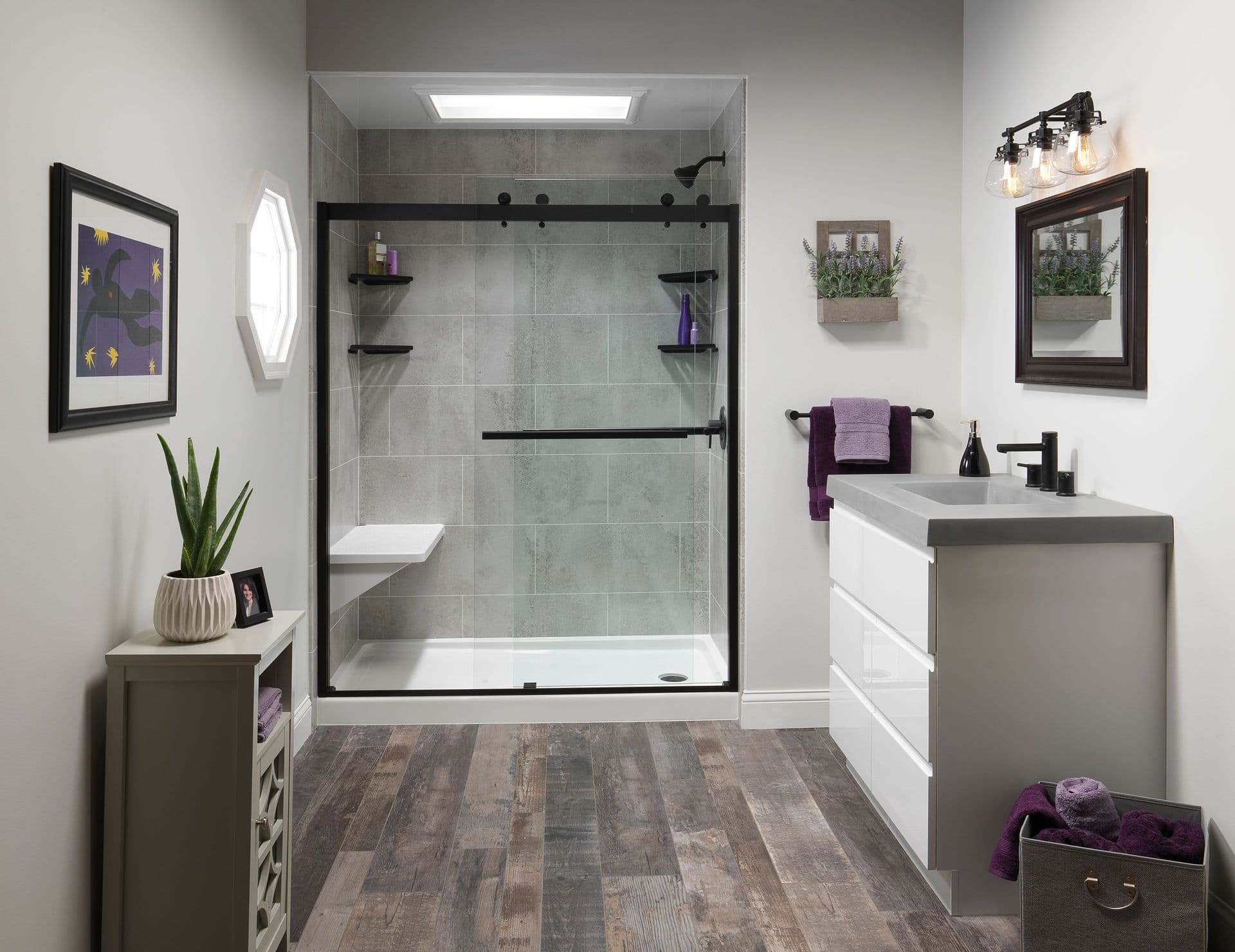 Modern bathroom with gray tile shower, black frame, and shelves. White vanity with gray countertop, plants, purple towels, and wood floor. Cozy vibe.