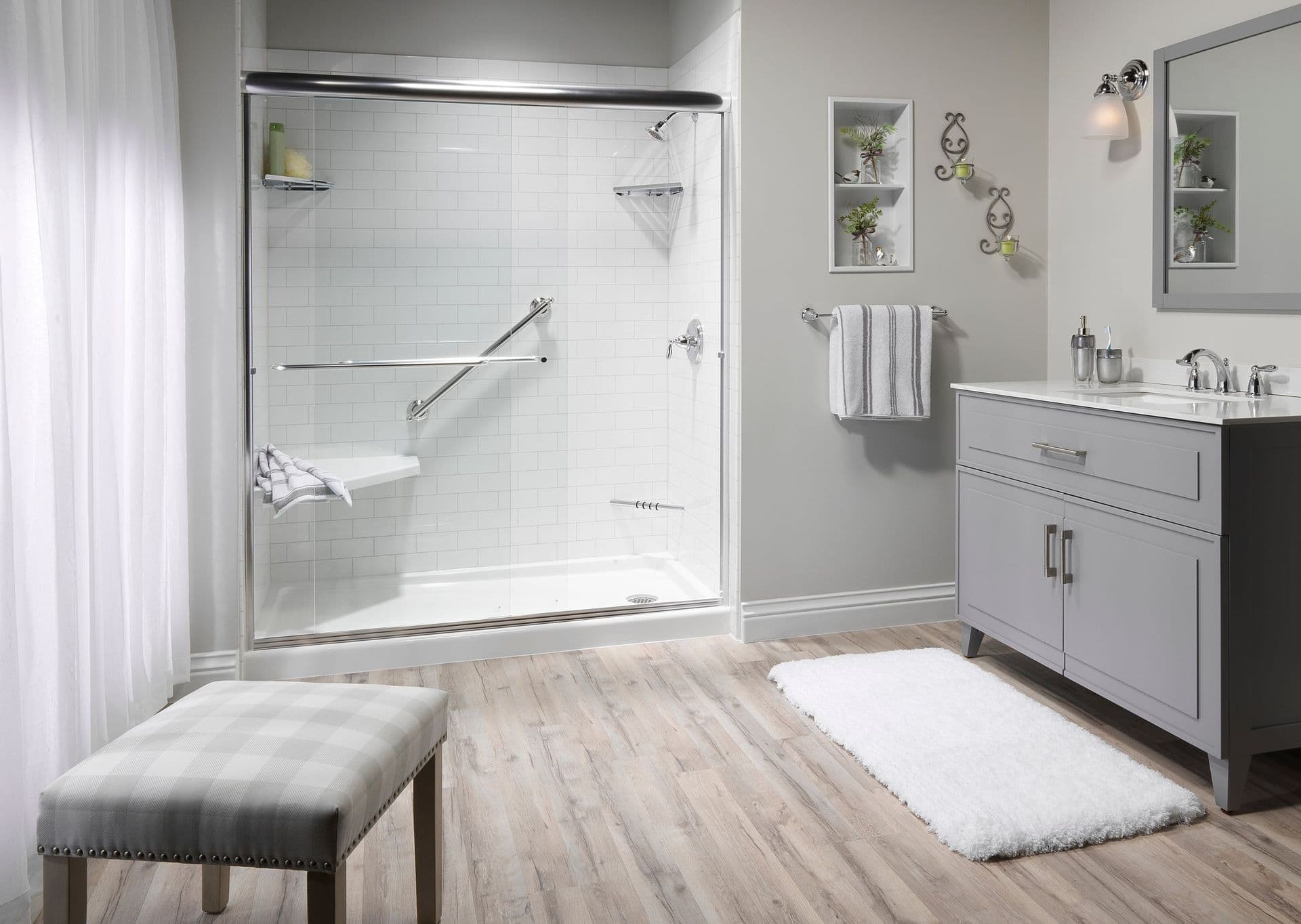 Bright, modern bathroom with a walk-in shower featuring a glass door, grab bars, and a bench. Light gray vanity, fluffy rug, and wooden floor. Calm ambiance.